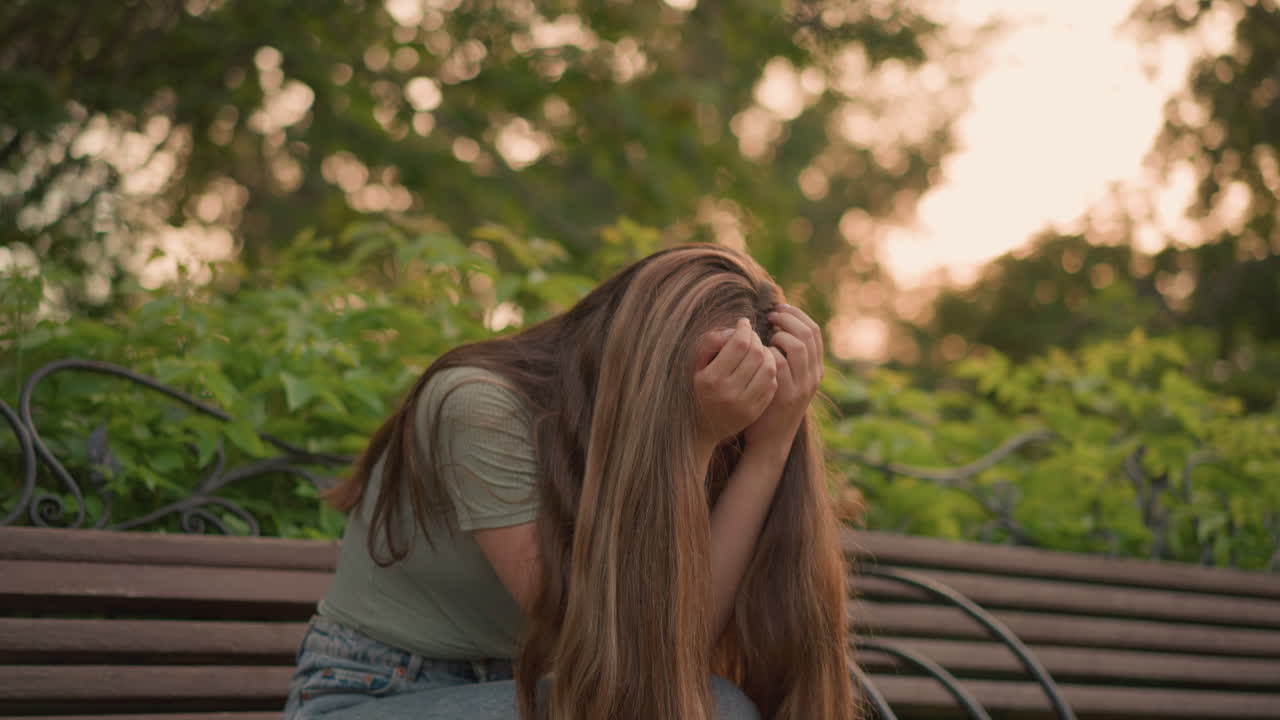 나무 벤치에 앉아있는 여성은 반사적이고 우울한 자세로, 손으로 얼굴을 고, 그녀의 긴 머리카락은 어깨 아래로 떨어지고, 따뜻한 빛은 나무와 울창한 초록색을 통해 필터링합니다.