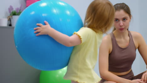 playful toddler holds large blue balloon while mother squats nearby, smiling warmly, gym background features colorful fitness equipment, adding to the cheerful and interactive atmosphere