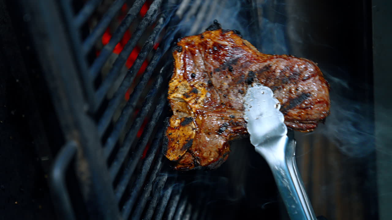 Piece of beef on the bone is cooked on the grill. Meat is turned with the tongs. Close up. Vertical view.
