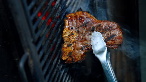 piece of beef on the bone is cooked on the grill. meat is turned with the tongs. close up. vertical view.