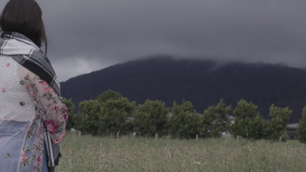 A young Hispanic woman stands pensively in a lush mountainous field under dramatic cloudy skies. The serene scene captures her thoughtful demeanor and contrasts with the moody backdrop