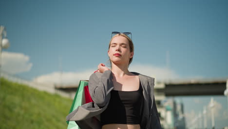 mujer blanca lleva la bolsa de compras hacia atrás mientras mira enfocado, el fondo muestra un puente superior y postes de farolas en un día soleado, cielos azules claros y vegetación