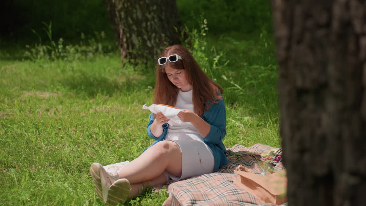 Close up of textured tree trunk with sunlight highlighting rough bark surface while blurred woman sits in background sewing cloth beside bag surrounded by green grass