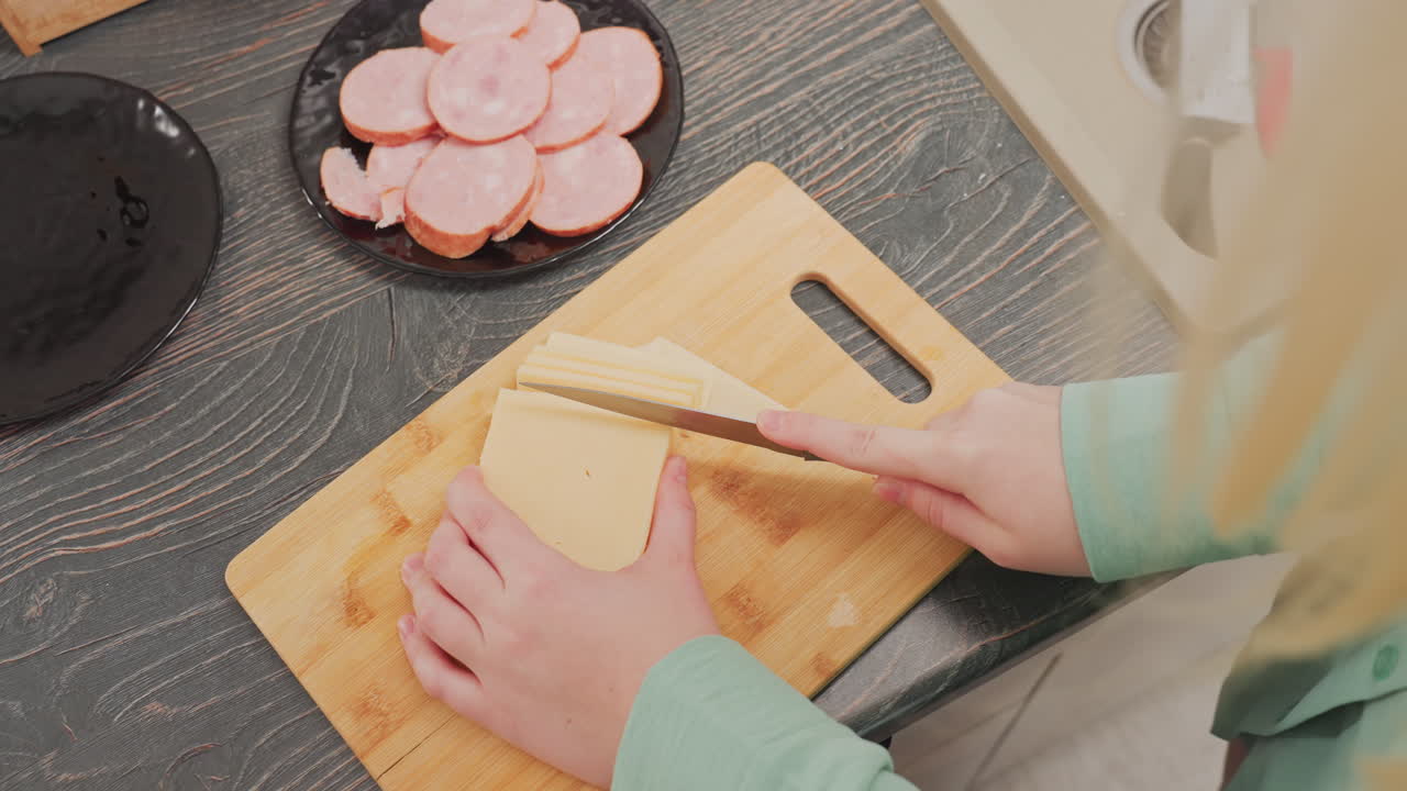 aerial view of woman in green shirt slicing block butter on wooden cutting board using knife in kitchen with black plate of sliced sausage in background on dark wood countertop