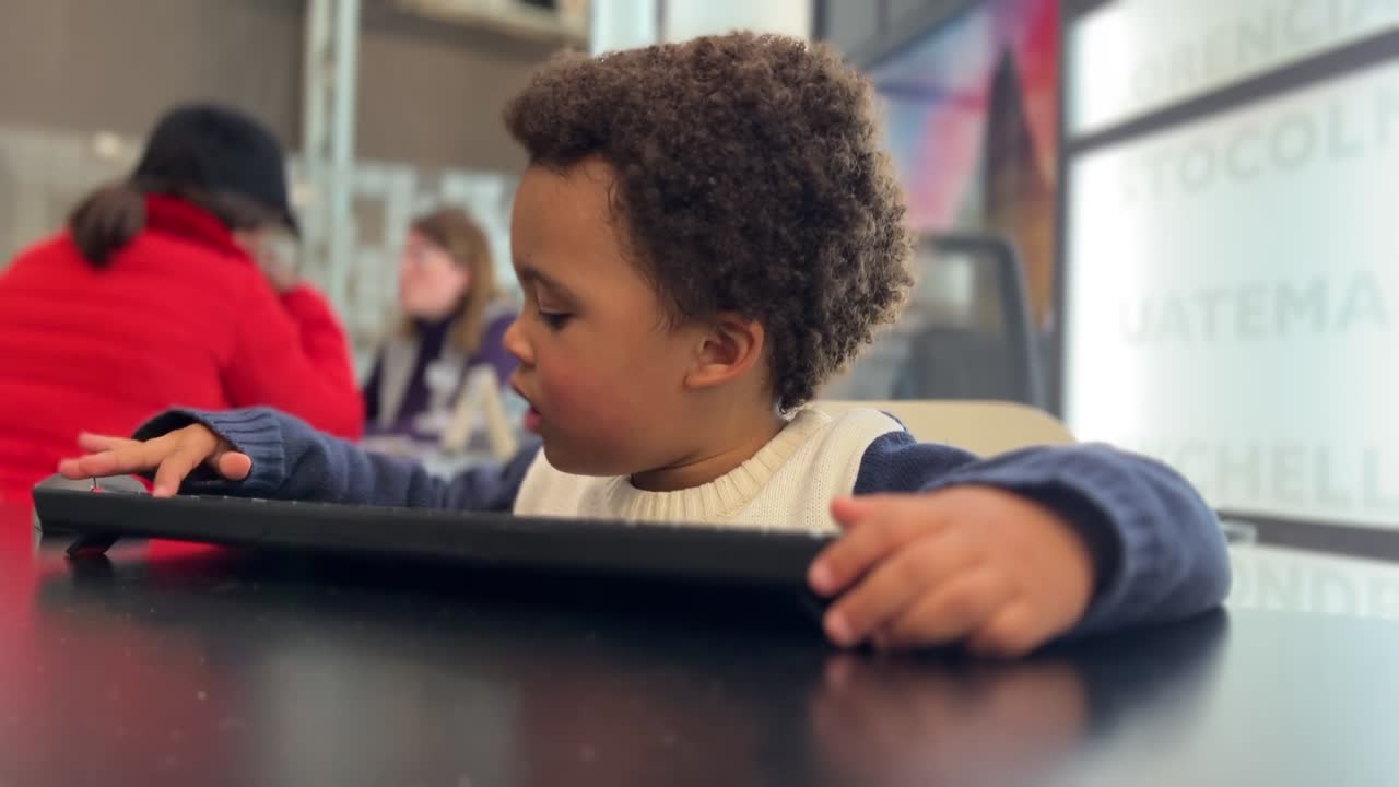 Bambino piccolo in ufficio: bambino esotico gioca alla scrivania della mamma, digitando come un professionista