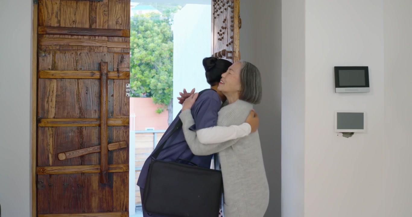 Assistente familiare donna di etnia diversa in divisa che fa visita, l'anziana che abbraccia, si tengono per mano, chiudendo la porta.