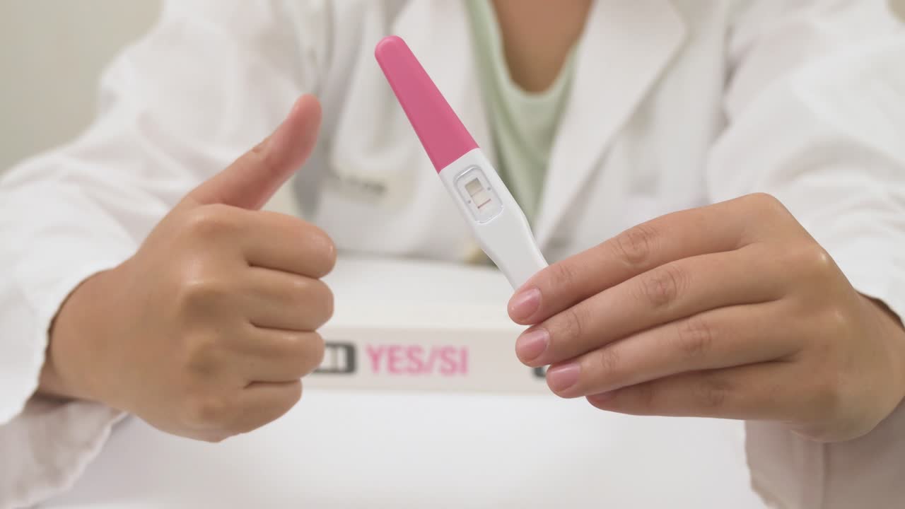 Female doctor holding a positive pregnancy test and gesturing thumbs up in a medical clinic