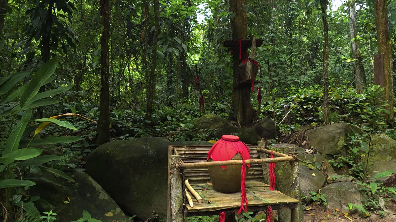 A traditional large red pot used for ceremonial and ritual purposes decorated with ribbons sits on a rustic wooden platform in a forest setting in Mari Mari cultural village, Kota Kinabalu, Malaysia