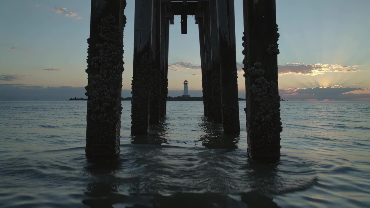 Un'onda che si infrange spruzza acqua, incorniciando i pali del molo ricoperti di cirripedi e rivelando un faro in lontananza all'alba