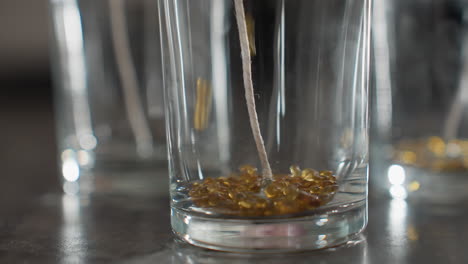 Close up of clear glass cup with white wick centered inside as brown decorative beads are being poured into it for candle or craft preparation, with surrounding cups softly blurred in background