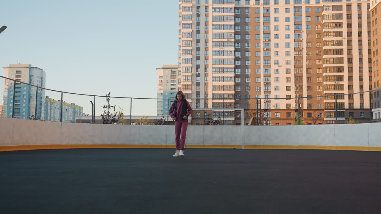 Female athlete jogging around outdoor court surrounded by tall apartment buildings, wearing purple tracksuit and white sneakers, stretching arms above head under soft evening sun