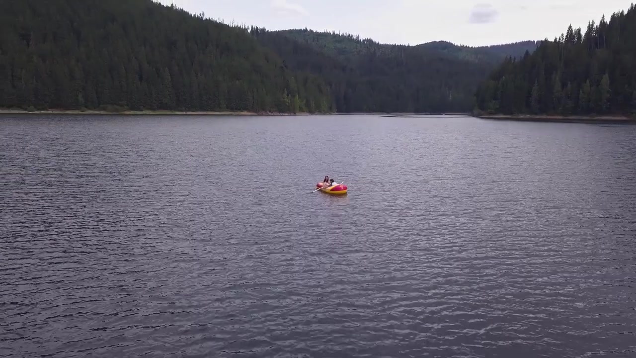 Un gommone gonfiabile con due persone alla deriva su un lago tranquillo, circondato da fitte colline boscose. Ideale per documentari sulla natura o contenuti di viaggio.