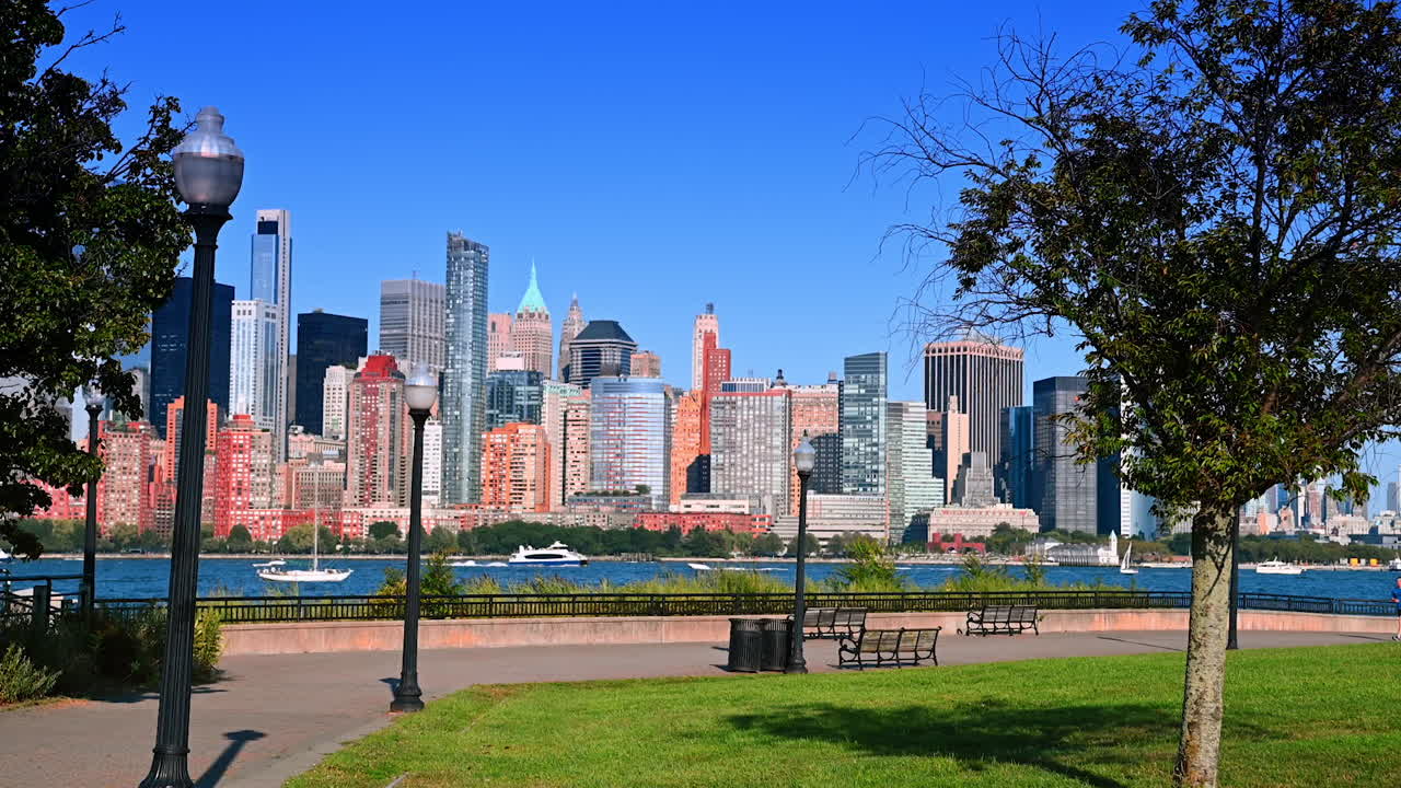 Park view of city skyline. Tall buildings rise against a clear blue sky, with a park in the foreground and boats on the water in the distance