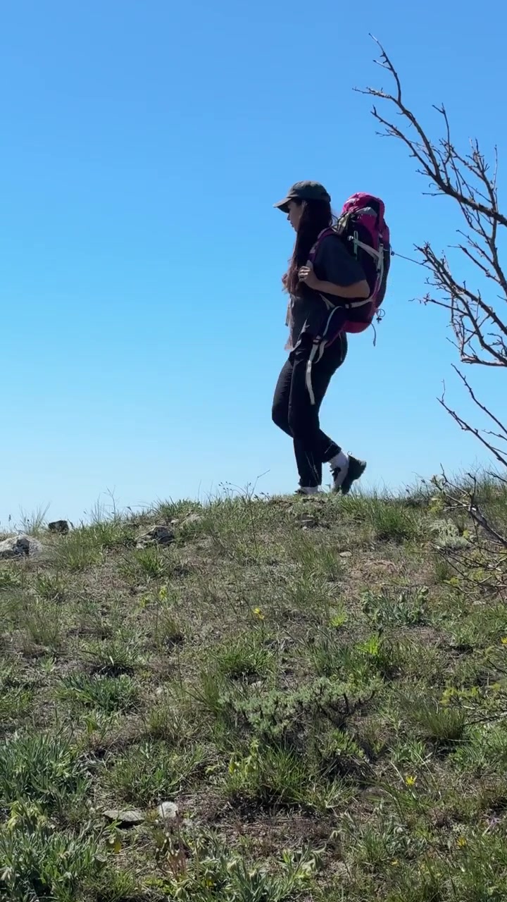 Una donna con uno zaino cammina in salita su un pendio erboso sotto un cielo azzurro limpido a Sudak, Crimea, Russia.