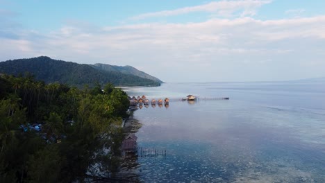 Scenic-aerial-view-rising-over-waterfront-houses-and-huts-overlooking-ocean-on-a-tropical-island-in-Raja-Ampat,-West-Papua,-Indonesia