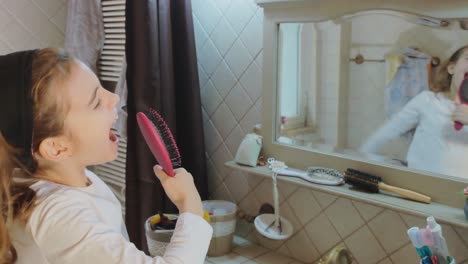 joyful young girl sings into hairbrush, pretending it's a microphone, in bathroom mirror