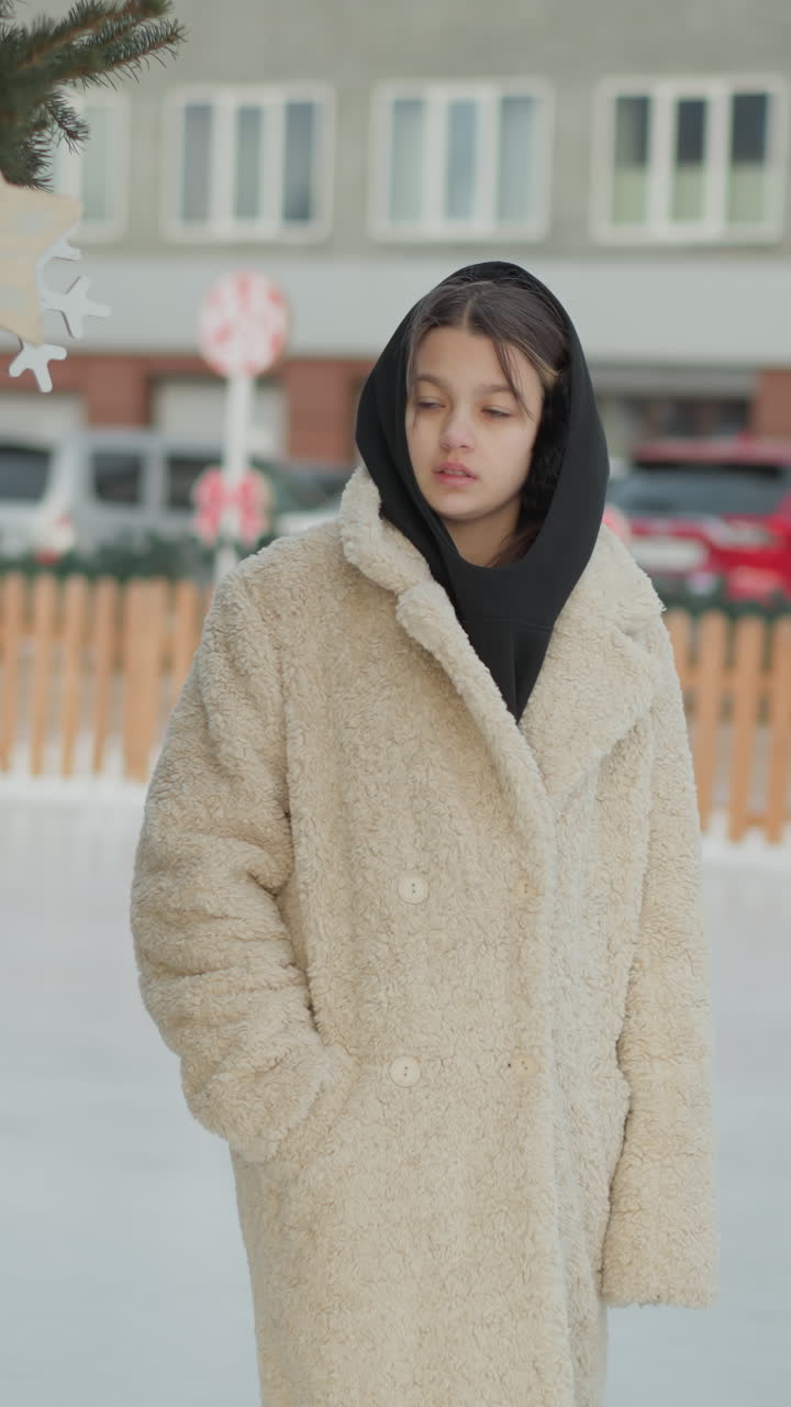 시어링 코트를 입고 야외 아이스 링크 옆에 서 있는 백인 십 대, 배경에는 도시 상점가와 막대 사탕 장식, 주머니에 손을 넣고 생각에 잠긴 시선, 눈발, 차가운 팔레트, 평온한 휴일