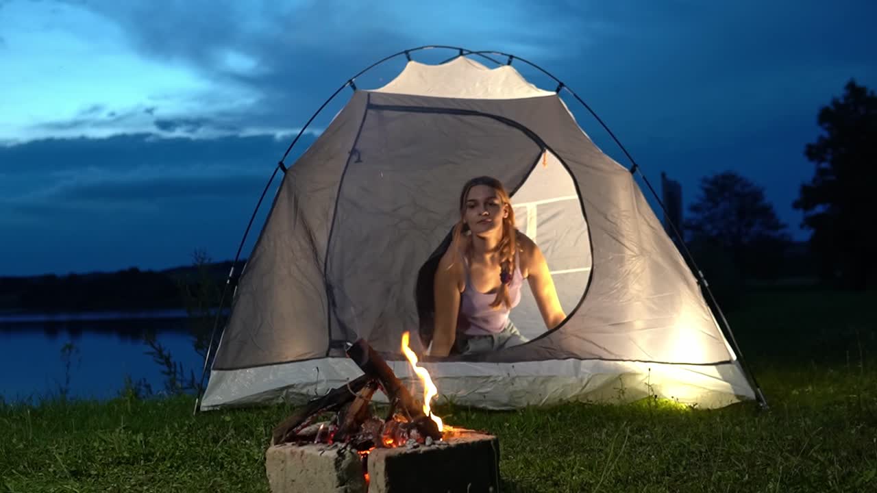 Kvinde åbner telt og kigger ud mod bålet nær en rolig sø om natten, der fanger hyggelig campingstemning og fredelig udendørs livsstil i Savci