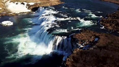 an aeriel shot of rainbow cast over faxi waterfall from a river that cuts through the snow covered countryside in iceland