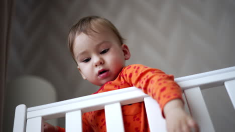 adorable baby boy standing in his cot looks at camera from top. lovely kid notices something aside, smiles adorably and hurries there. low angle view.