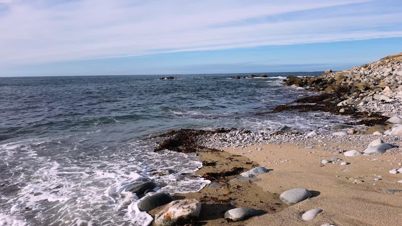 Waves with slight foam gently lapping a pebble and sandy beach with rocks out to sea on a bright calm day