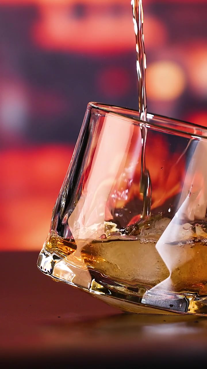 Un flusso di whisky dorato riempie un bicchiere dondolante unico con ghiaccio, su uno sfondo bokeh caldo.