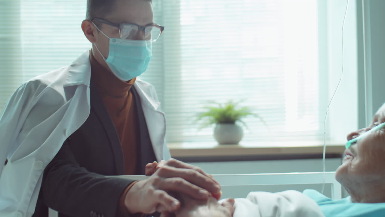Man in Mask Supporting Senior Mother in Hospital Ward
