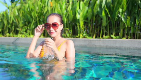 an attractive woman, chest high in a swimming pool, looks at the camera as she adjusts her sunglasses