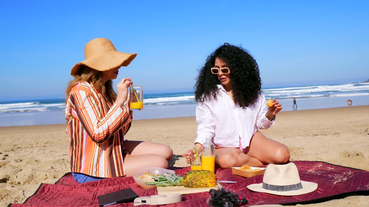 To glade kvinner nyter en piknik og drikke på en solrik strand