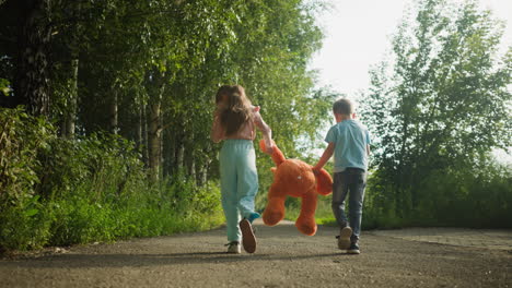 two siblings standing on sunny forest path as older girl holds large orange teddy bear and younger boy crouches down to fix shoe, surrounded by lush greenery and tall birch trees