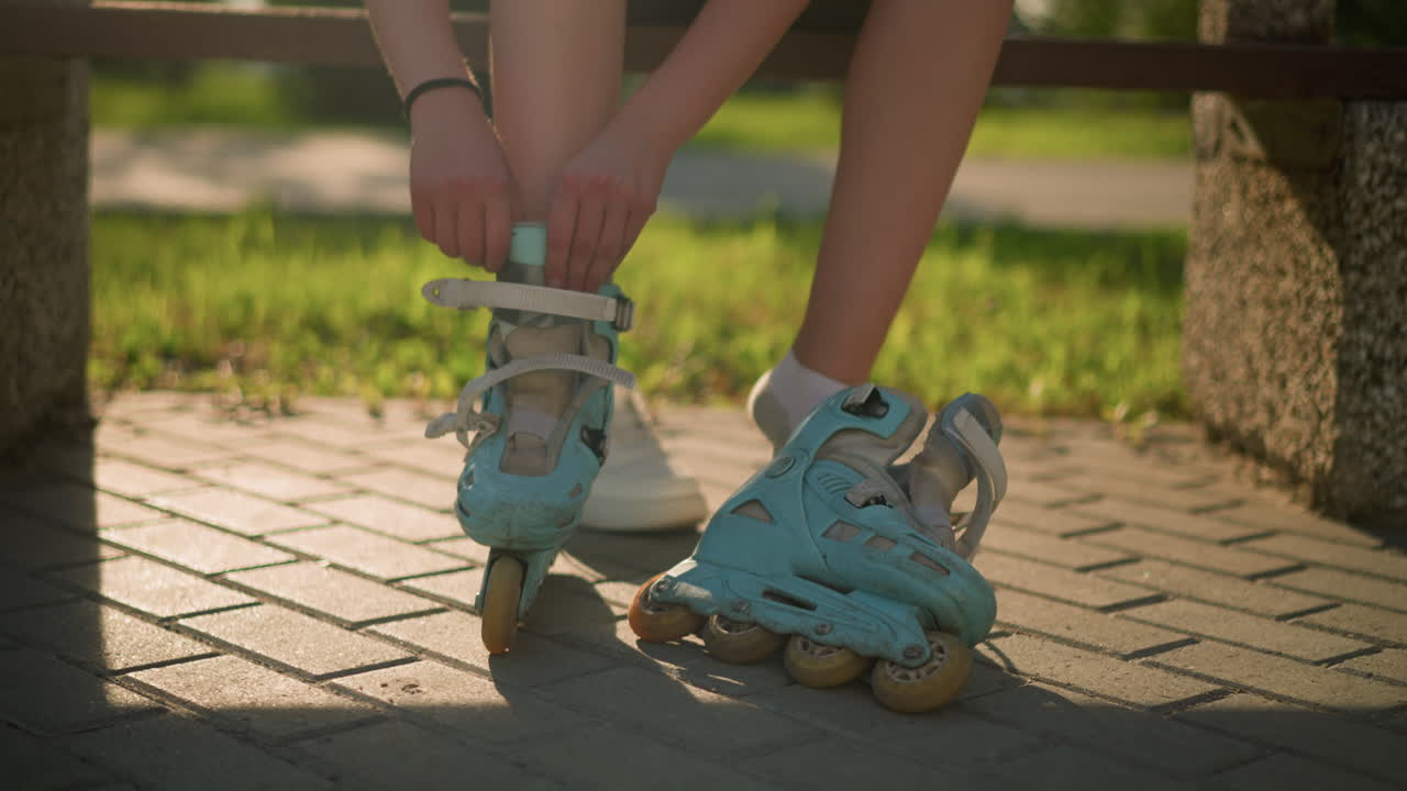 Leg view of individual adjusting strap on cyan roller skates while sitting on bench, wearing black bangle on right hand in background, warm sunlight casts shadows with greenery behind