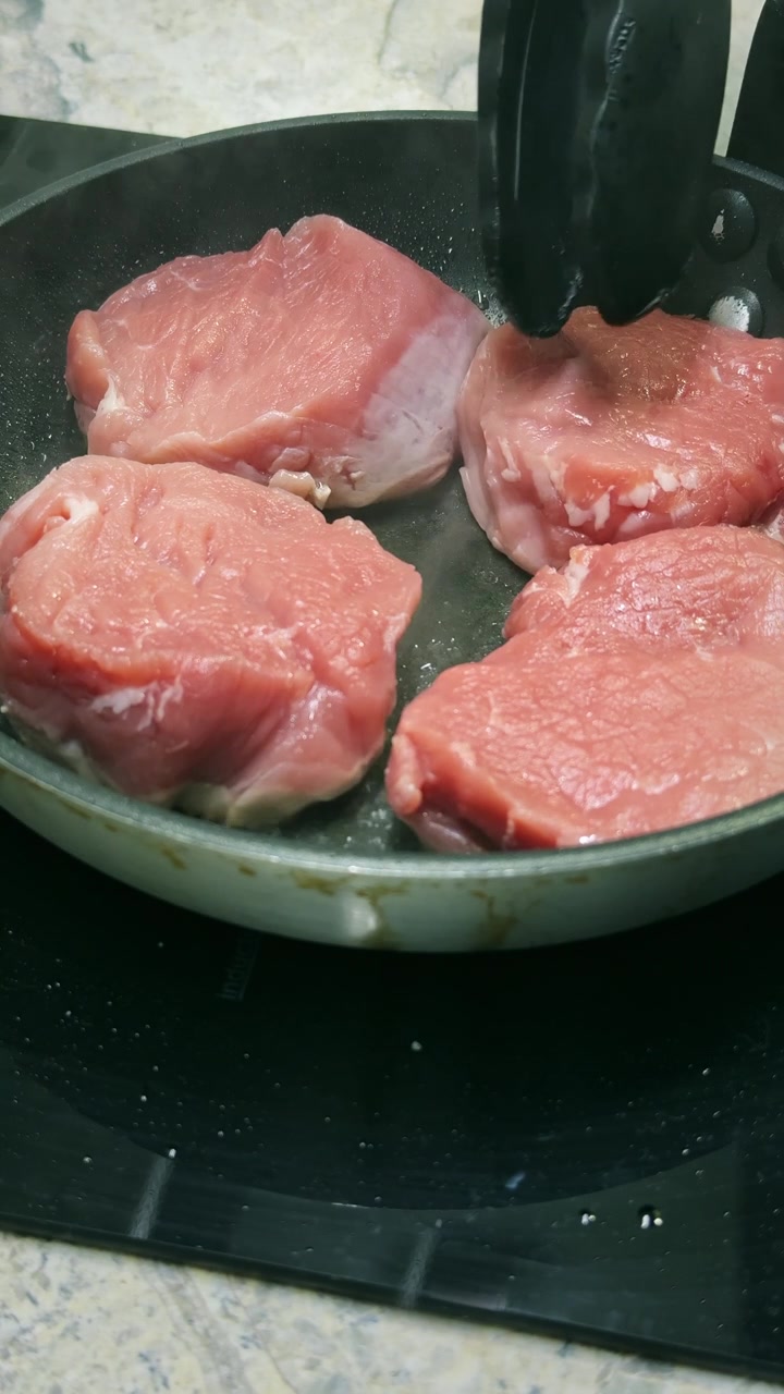 Close-up seorang koki menempatkan medali tenderloin babi mentah ke dalam minyak panas di wajan. Rekaman memasak 4K vertikal yang menangkap awal proses pemanggangan cepat.