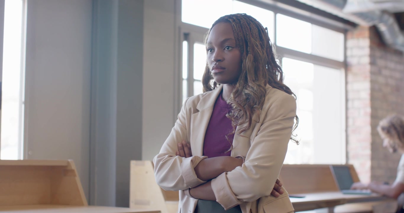 La telecamera si avvicina a una donna afroamericana con blazer beige a braccia conserte, che osserva uno studente al laptop.