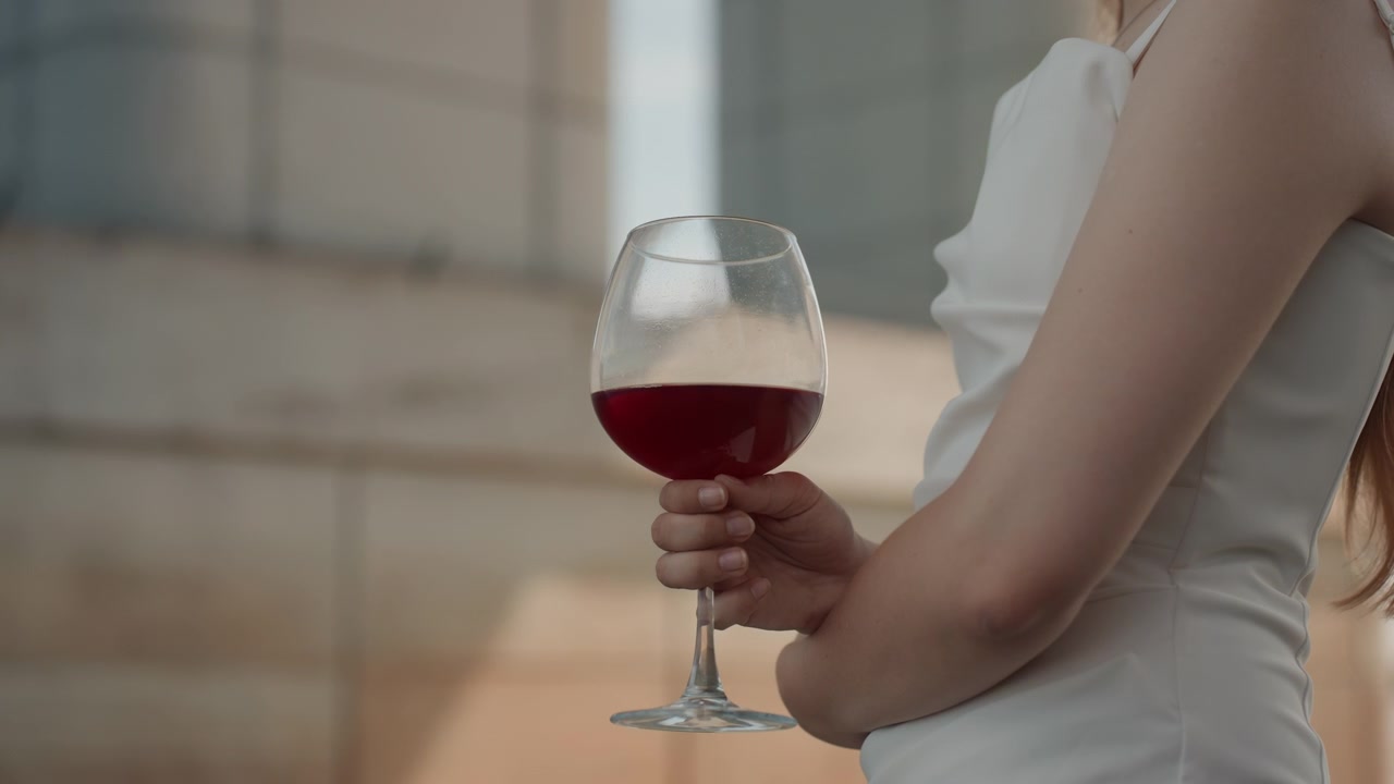 Donna in abito bianco tiene un bicchiere di vino rosso, con luce naturale soffusa e uno sfondo urbano esterno. La scena è calma e composta, concentrandosi sul braccio e sulla mano del soggetto che tiene la bevanda contro uno sfondo sfocato