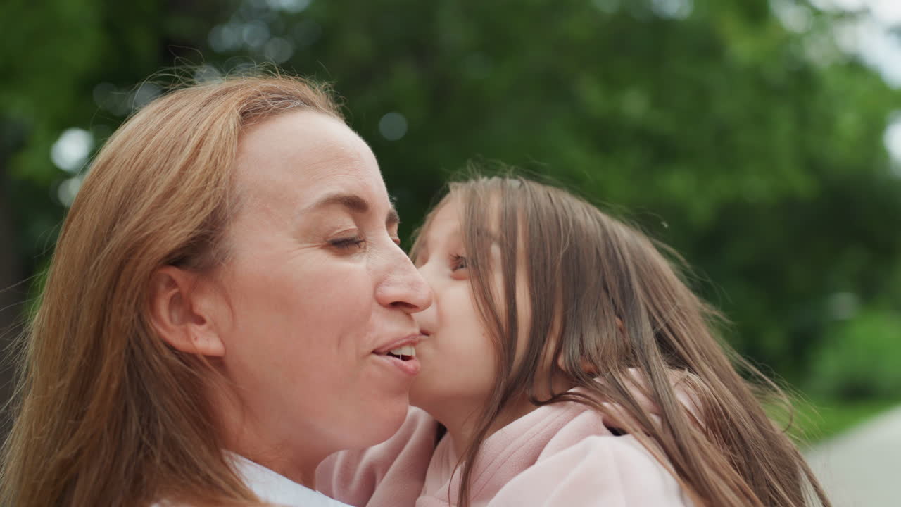 Mutter und Tochter umarmen sich im Park im Freien (Nahaufnahme), mit sanften Küssen und Lachen. Eine herzliche Umarmung, die Mutter hält das Kind mit pinker Jacke vor grünem Baumbestand. Ein aufrichtiger, freudiger Moment inniger Verbundenheit und fürsorglicher Beziehung.