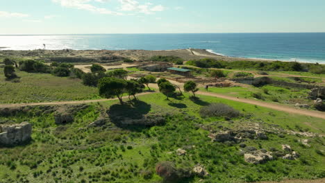 An-aerial-view-over-the-historical-site-of-the-Tombs-of-the-Kings-in-Paphos,-with-its-ancient-ruins-amidst-a-green-landscape,-overlooking-the-Mediterranean-Sea-under-a-clear-blue-sky