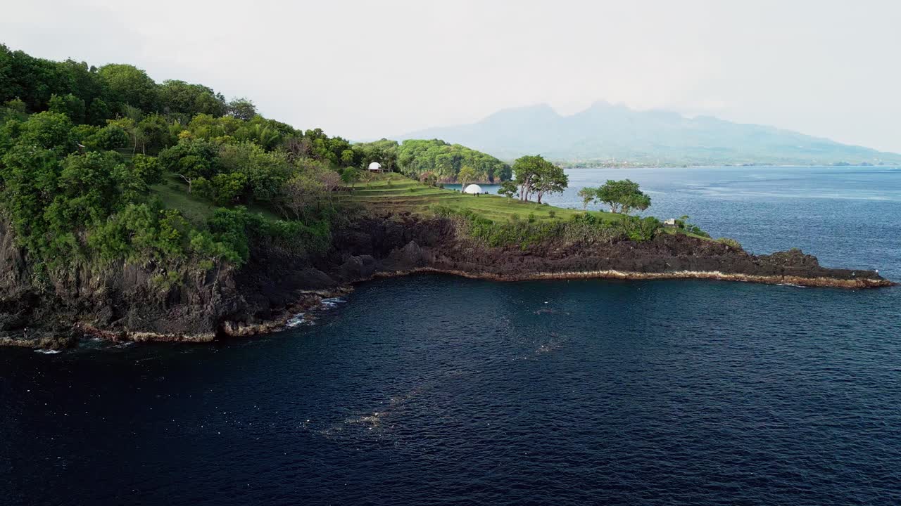 Un filmato aereo rivela un campeggio su un'isola tropicale con moderne tende a cupola, posizionato su una robusta roccia vulcanica, con vista su acque oceaniche calme e montagne distanti all'orizzonte