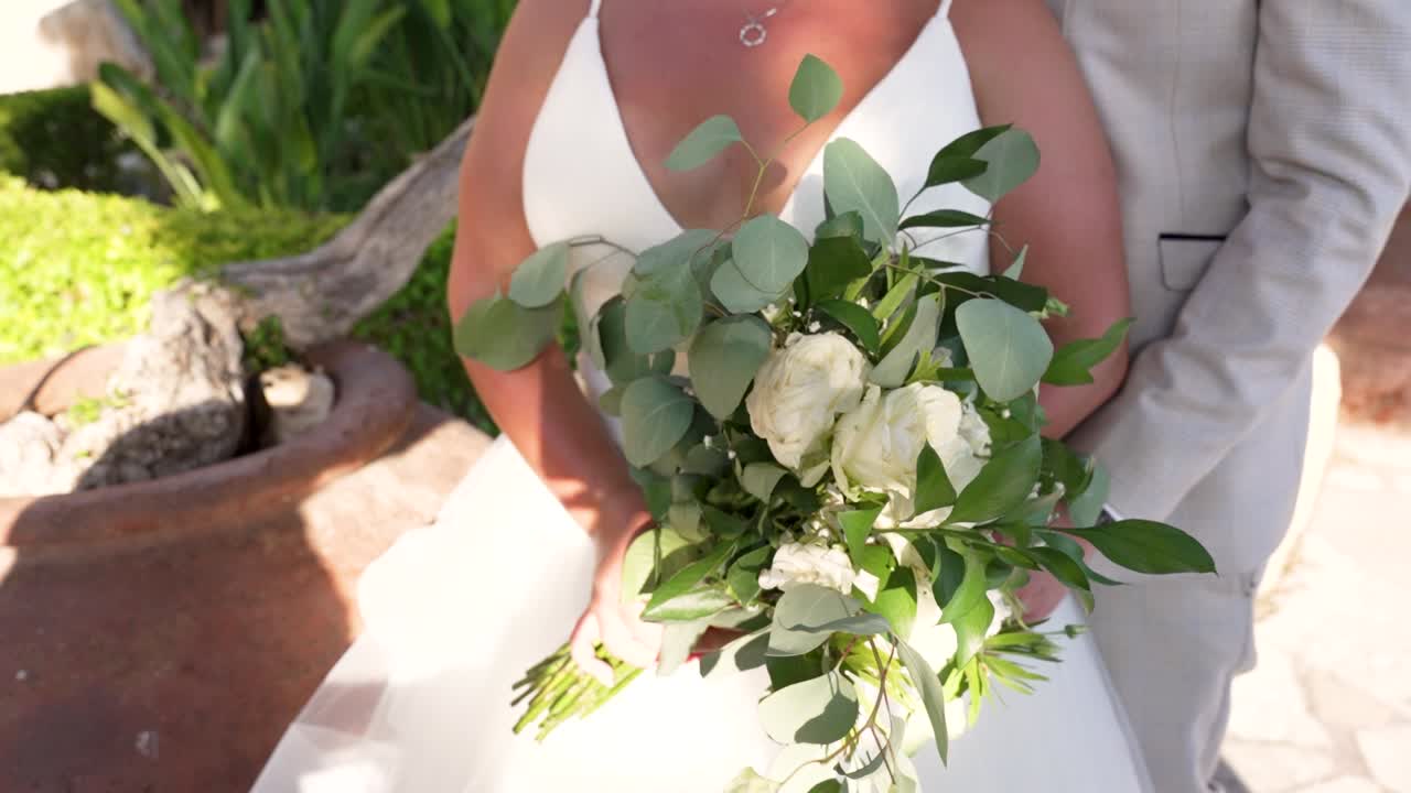 A radiant bride stands gracefully outdoors, holding a bouquet filled with elegant white roses and lush greenery. The joy of the moment is evident as she prepares for the special day