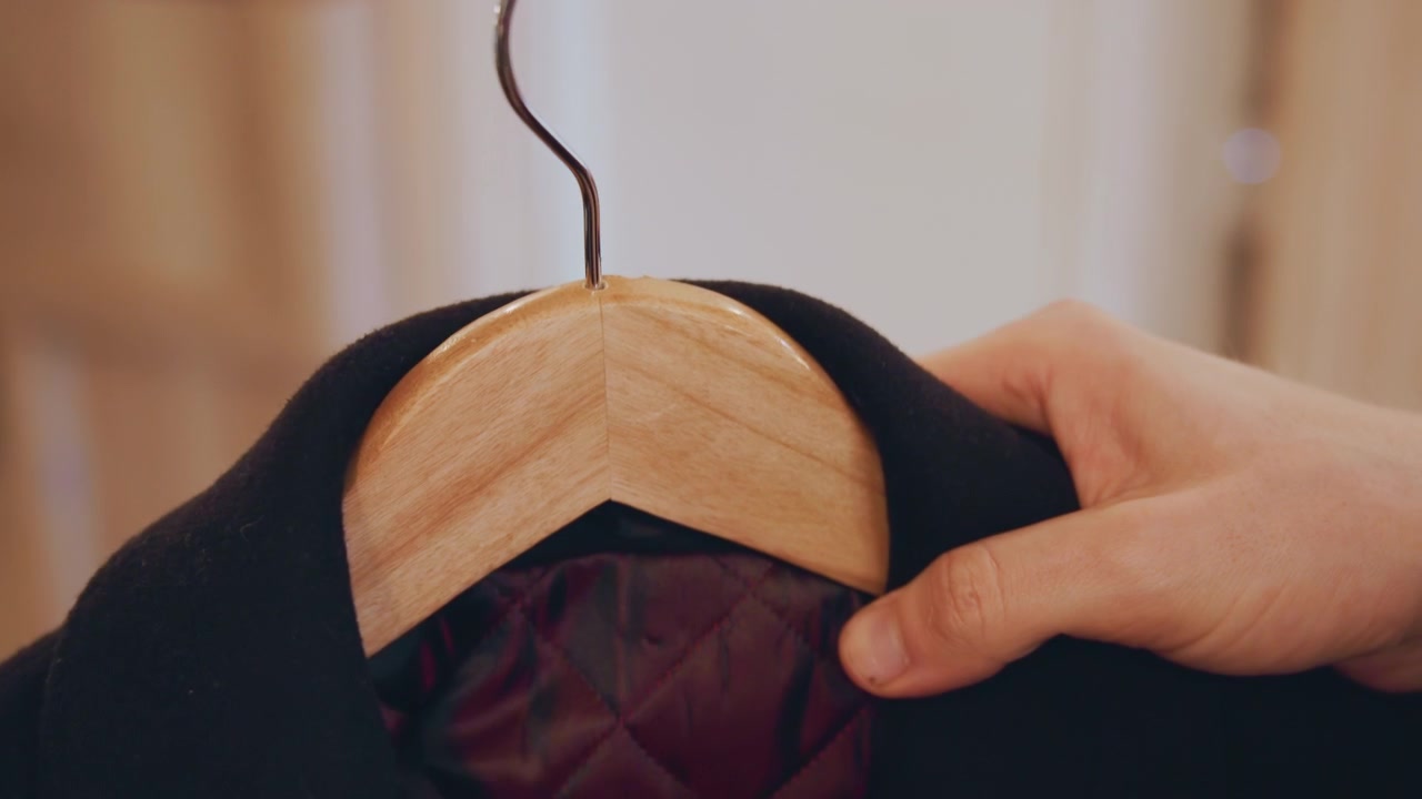 Primo piano di mani che posizionano un indumento nero su appendini di legno. La persona sistema il tessuto su più ganci assicurando un posizionamento corretto durante tutta la sequenza. Una luce interna soffusa evidenzia la texture.