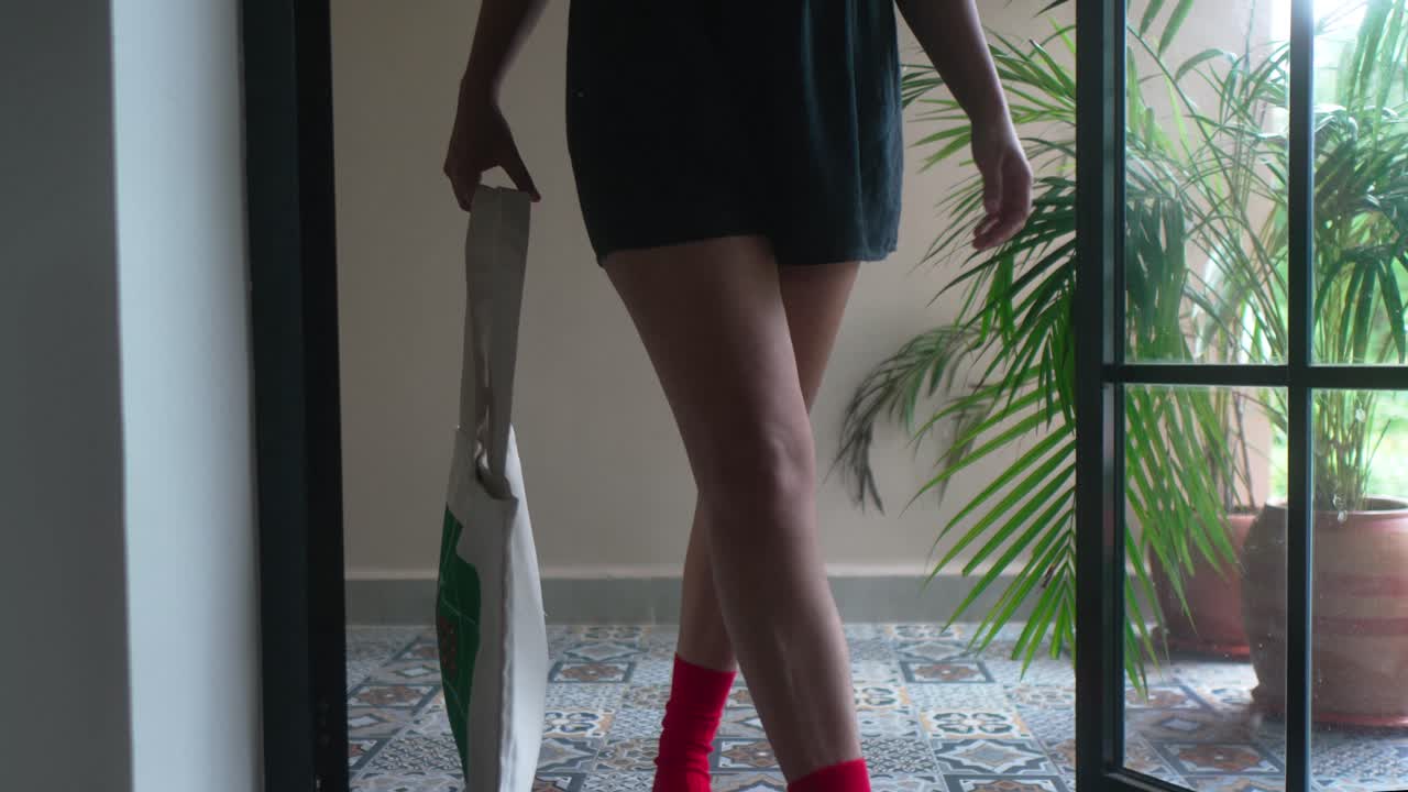 Donna che indossa un abito nero e stivali rossi sorprendenti, con una tote bag, che cammina da una stanza con piastrelle a fantasia e una pianta verde, esprimendo moda e stile di vita moderni.