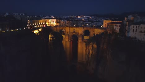 drone captures iconic puente nuevo lit at night with glowing street, cliffside house, and wide view of historic ronda townscape under dark sky in southern spain, creating a scenic night panorama