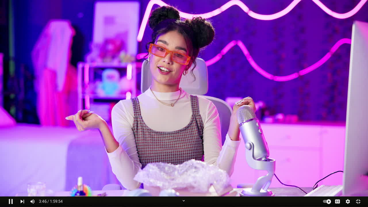Woman with orange glasses and hair buns in a neon lit bedroom with a microphone