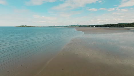 Aerial-shot-of-birds-flying-low-over-beach,-calm-mirrored-water-reflecting-blue-sky-and-clouds,-seagulls-fly-over-sandy-beach-at-low-tide-on-Scottish-coast