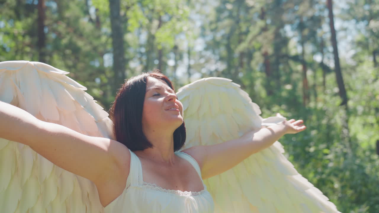 Elegant girl with angel wings smiles warmly under sunlight while turning around in green forest, arms open in joy and peace, expressing beauty, happiness, and connection with nature