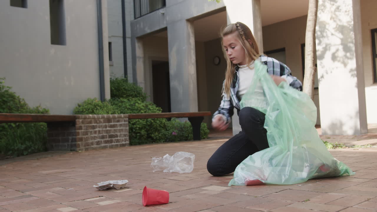 Young young girl cleaning up litter outdoors, collecting trash in green bag, at school