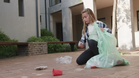 young young girl cleaning up litter outdoors, collecting trash in green bag, at school