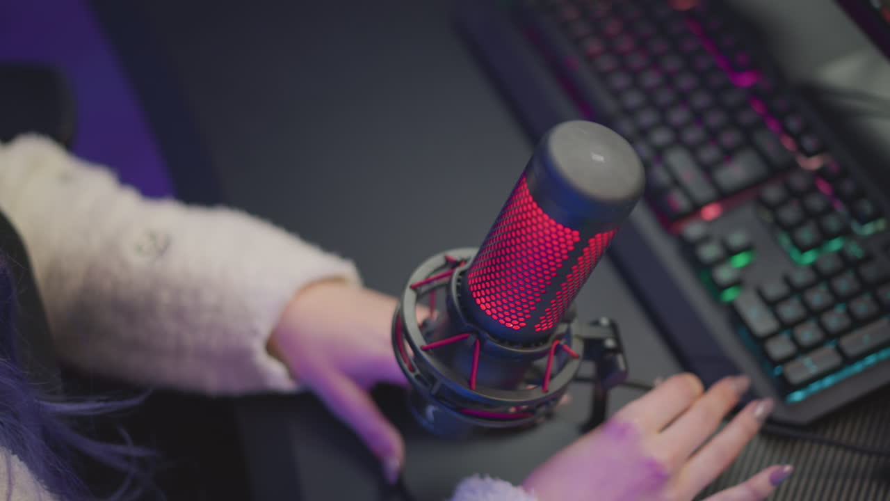lady in fluffy jacket adjusts microphone cable with one hand while touching rgb keyboard beside glowing desktop monitor displaying forest wallpaper in dim lit workspace with moody colored lights