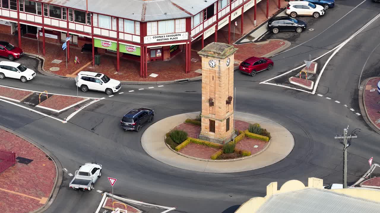 Vehicles drive through a roundabout with a central clock tower in Coonabarabran, Australia. Overhead view, steady daylight, urban residential setting