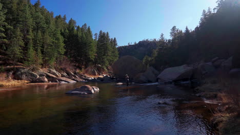 Cinematic-sunny-Colorado-river-fly-fish-fisherman-with-waders-using-rod-fishing-in-water-Cheesman-Canyon-Deckers-Conifer-Evergreen-late-winter-early-spring-shaded-mountainside-to-the-left-movement