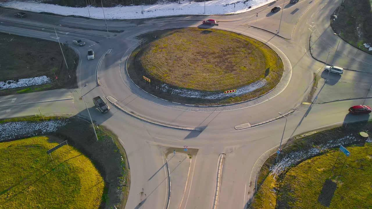 Aerial drone footage of cars driving on a roundabout in Lagri Estonia during a sunset or sunrise during a cold winter sunny day with white snow on the road side while cars and vehilces drive around.